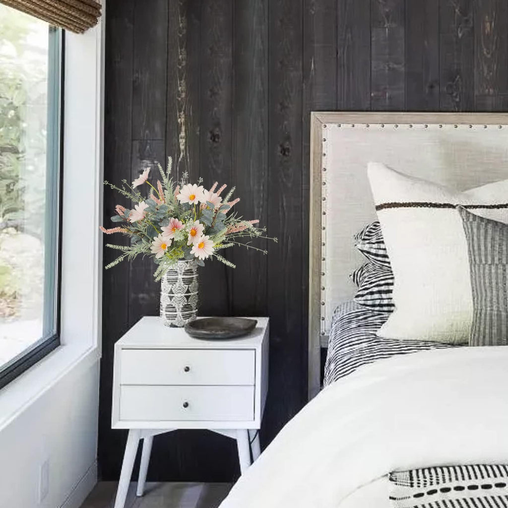 Artificial wildflower bouquet with pink daisies and eucalyptus in a patterned vase on a nightstand next to a bed.