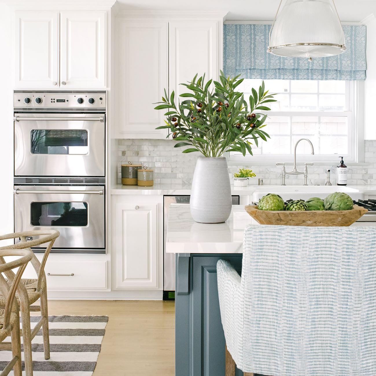 Artificial olive branches with leaves and olives in a vase on a kitchen island, with ovens and sink in background.