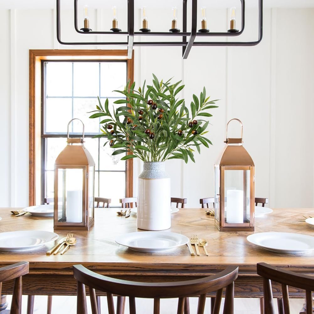 Artificial olive branches with leaves and fruits in a vase on a dining table with place settings and lanterns.