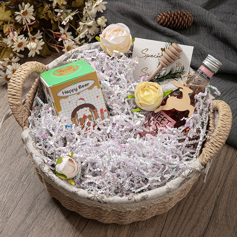 Gift basket filled with iridescent crinkle paper shred, a small box, a bottle, and decorative flowers.