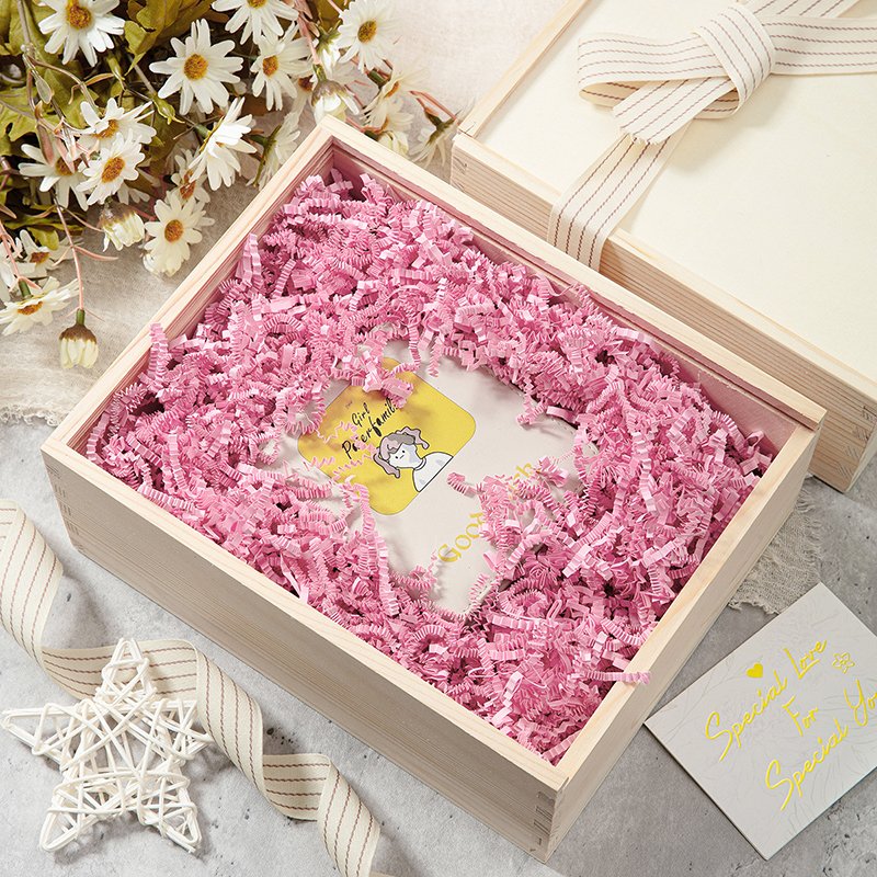 Open wooden gift box filled with pink crinkle paper shred and a card.
