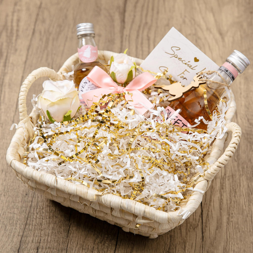 Gift basket filled with white and gold crinkle paper shred, two small bottles, and decorative roses.
