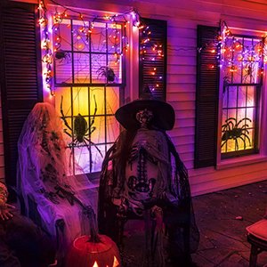 Halloween decorations with orange and purple string lights, spiders, skeletons, and a pumpkin on a porch.