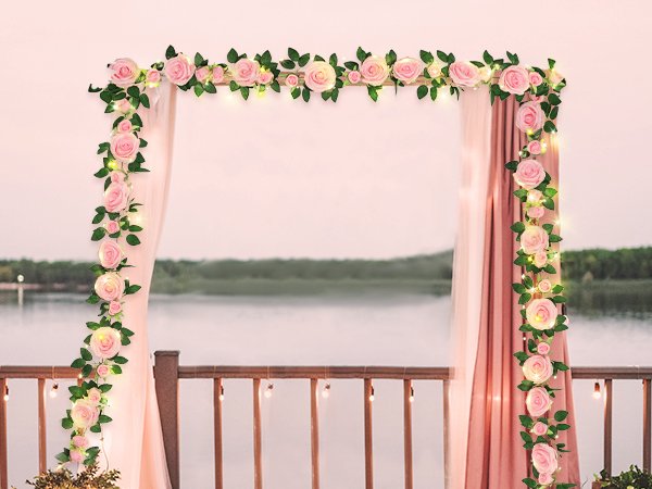 Pink artificial rose garland with fairy lights draped over a wedding arch with sheer pink curtains and a lake view