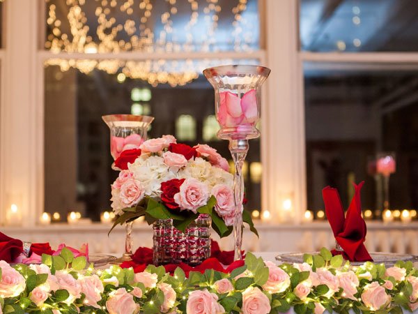 Pink and red roses with fairy lights draped along a table setting with candles and hurricane vases.