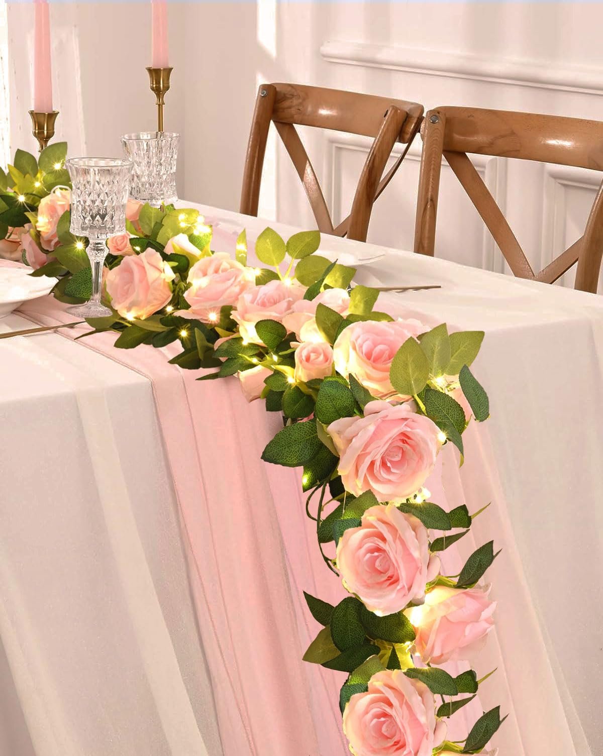 Pink artificial rose garland with fairy lights draped across a table setting with chairs in the background.