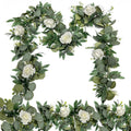 Artificial eucalyptus and white rose garland arranged in a decorative frame on a white background.