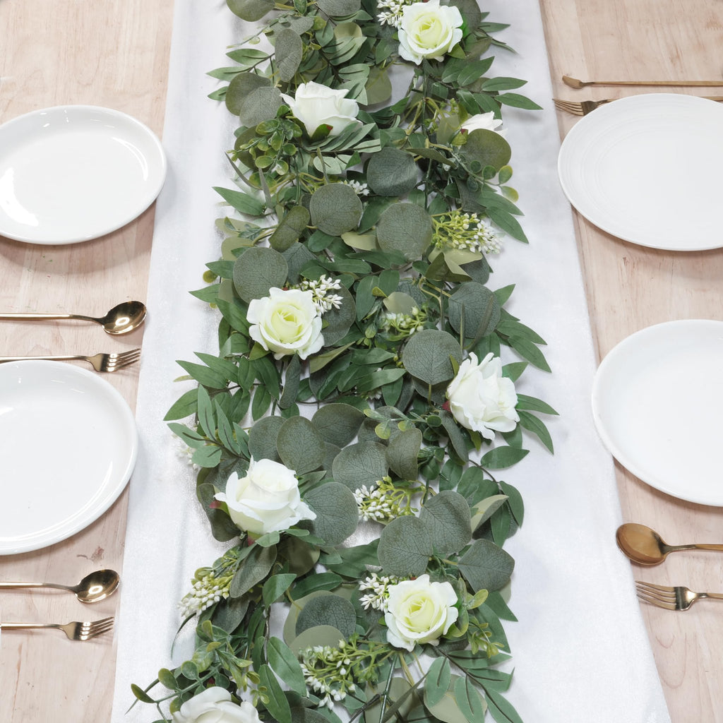 Artificial eucalyptus and white rose garland draped across a table setting with white plates and gold cutlery.