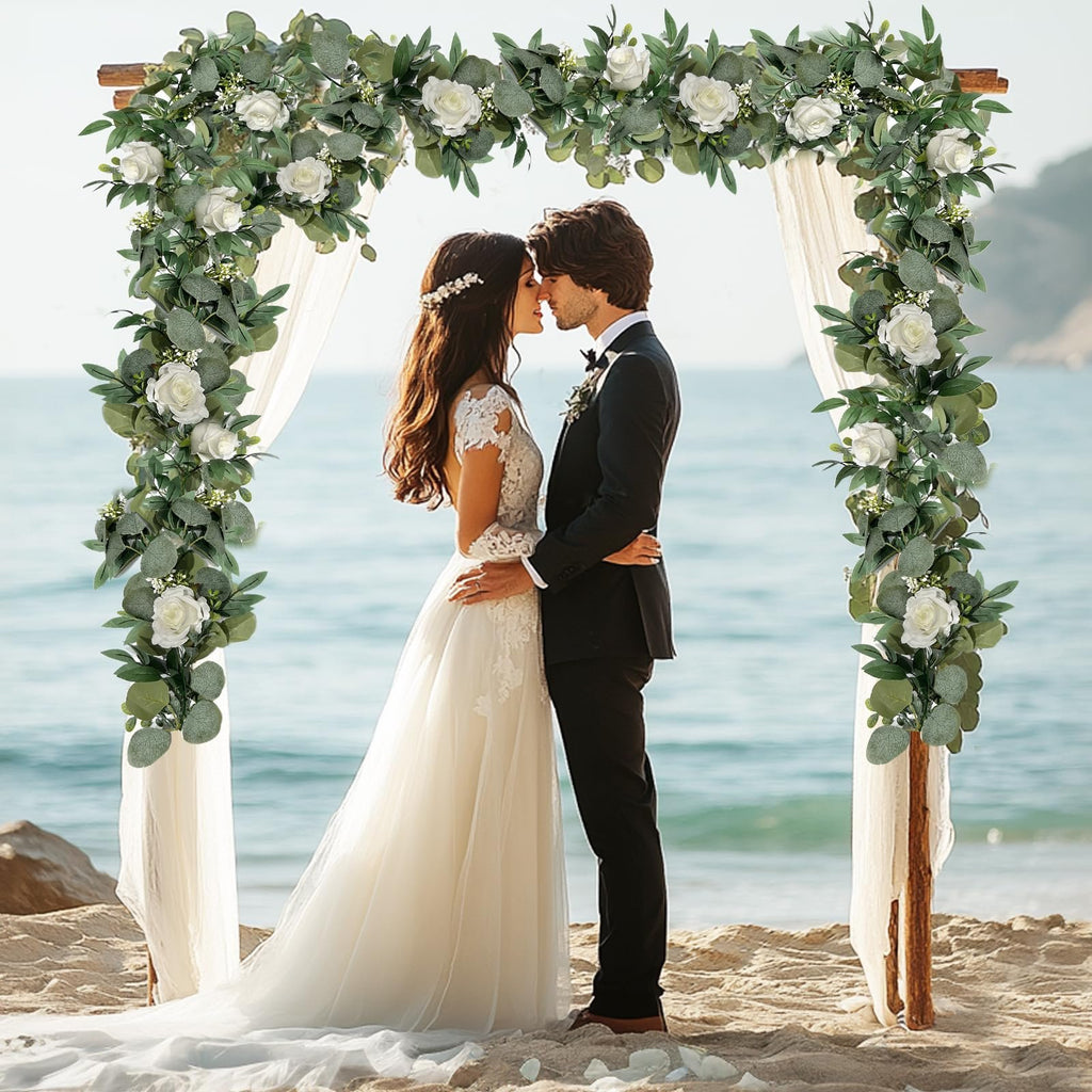 Wedding arch decorated with artificial eucalyptus and white roses on a beach with a couple kissing