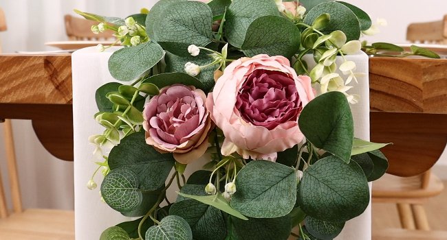 Artificial peony and rose garland with eucalyptus leaves draped over a white table runner on a wooden table.