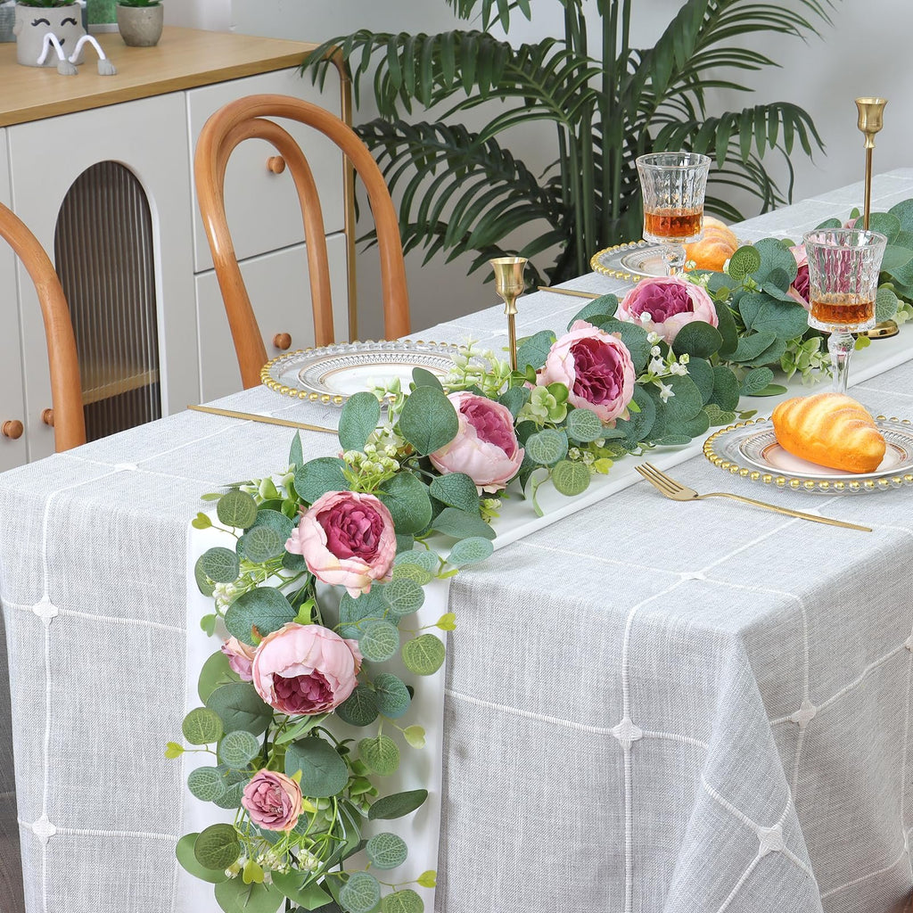 Artificial peony and eucalyptus garland draped across a table setting with plates, glasses, and bread.