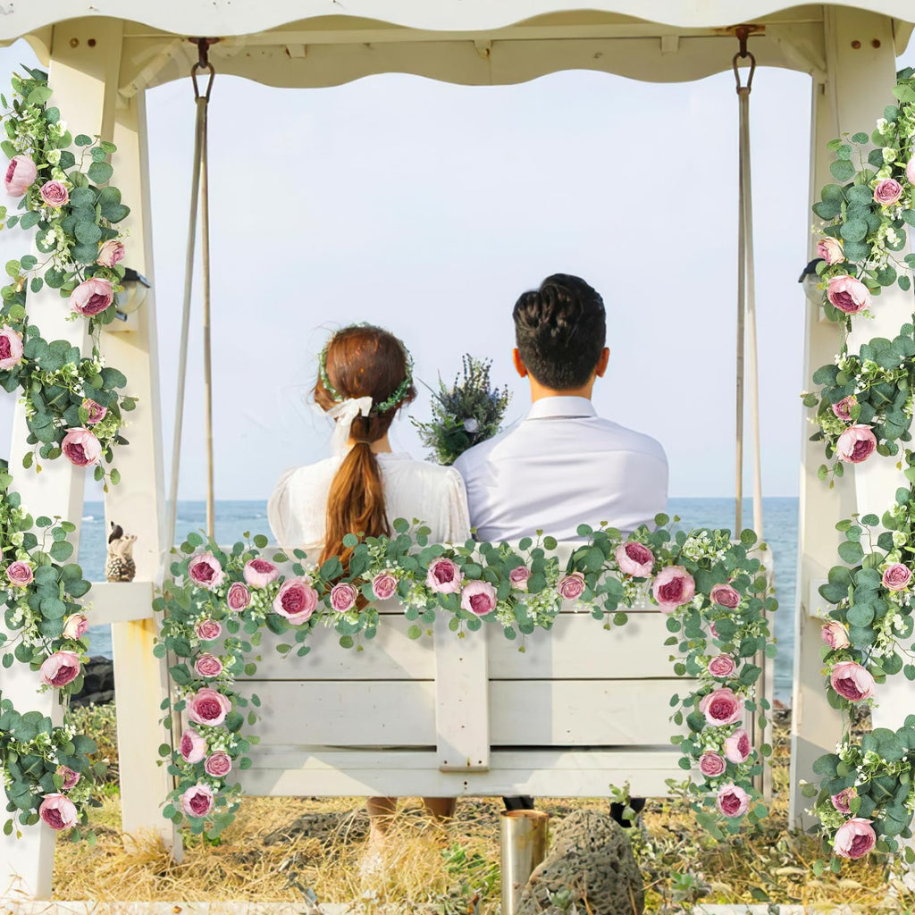 Artificial pink peony and eucalyptus garlands draped on a white swing with a couple sitting on it.