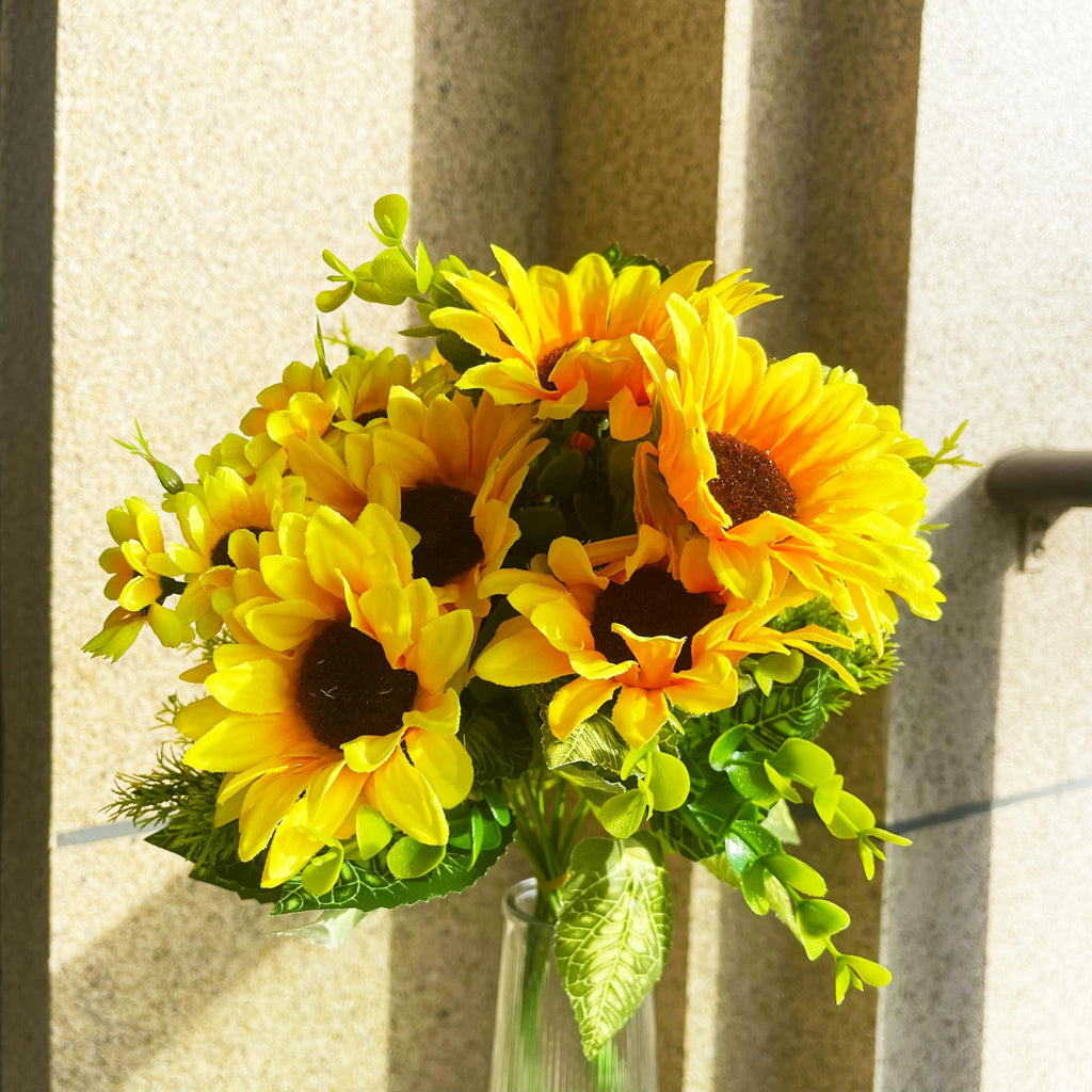 Bouquet of artificial sunflowers with eucalyptus and fern grass in a clear vase against a textured wall.