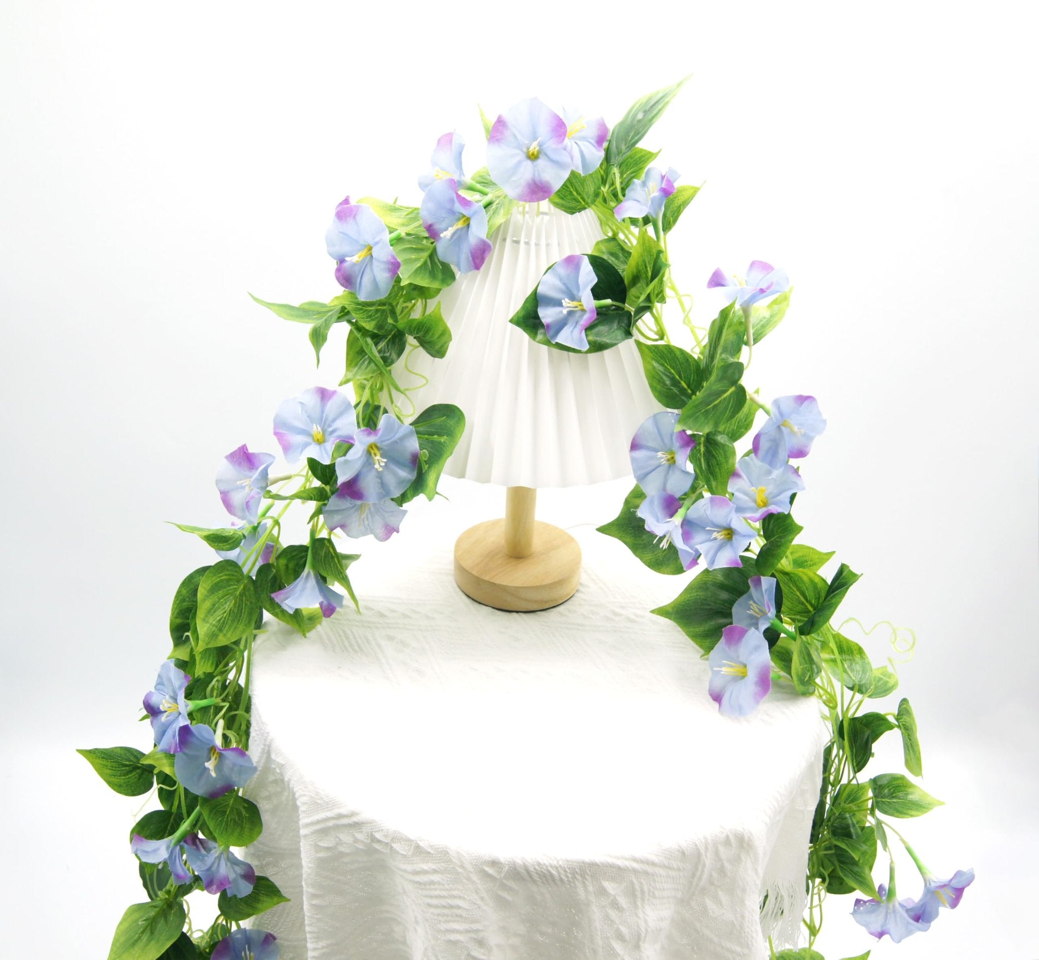Artificial morning glory vine garland with purple and blue flowers draped over a white pleated lampshade on a table.