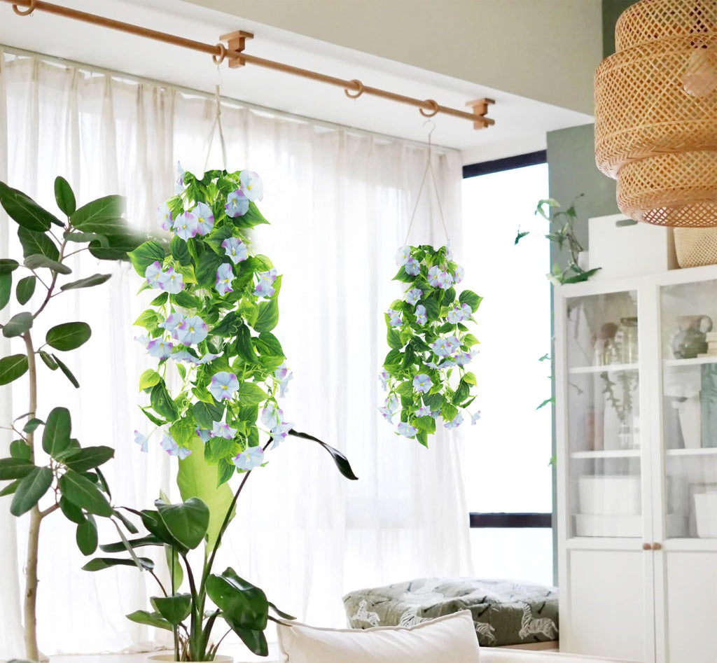 Two hanging artificial morning glory vine garlands with purple and white flowers and green leaves, displayed indoors near a window.