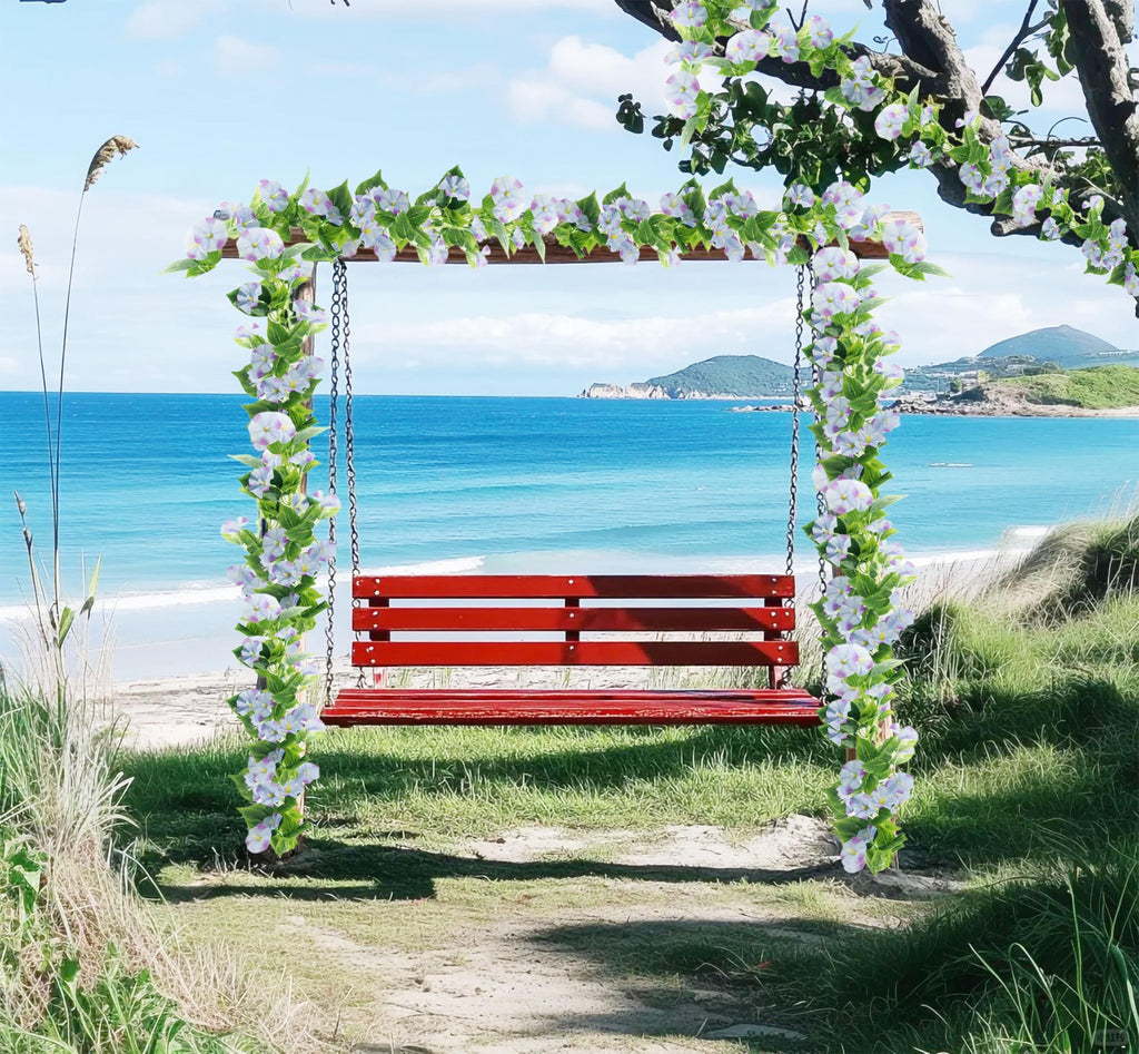 Red swing decorated with artificial morning glory vines and flowers on a beach with blue ocean and sky