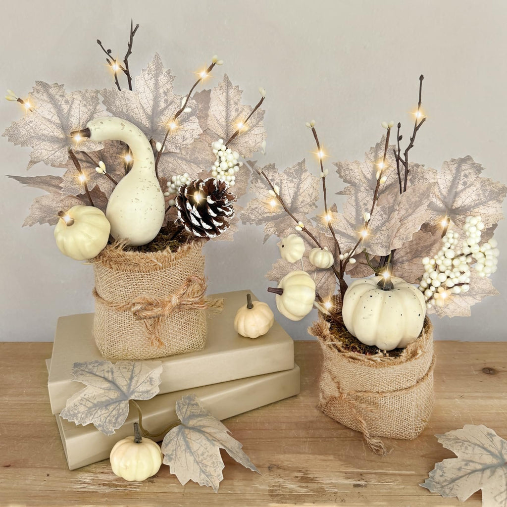 Two pre-lit artificial maple trees with pumpkins, pinecones, and berries in burlap pots on a wooden surface.