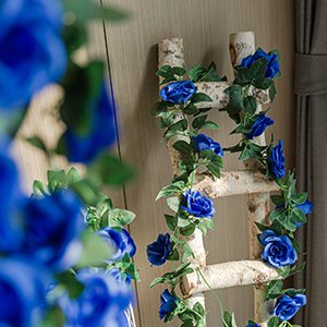 Artificial blue rose vine garland with green leaves draped on a birch wood ladder against a curtain.