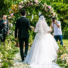 Wedding ceremony with a couple walking down an aisle under a floral arch, a photographer captures the moment.