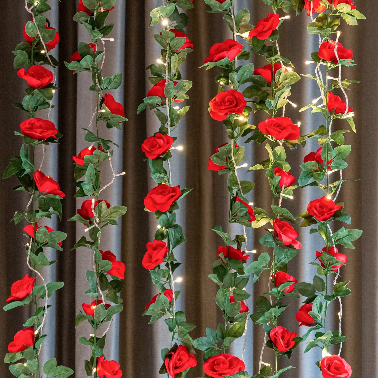 Artificial red rose vine garland with green leaves and fairy lights against a dark curtain backdrop
