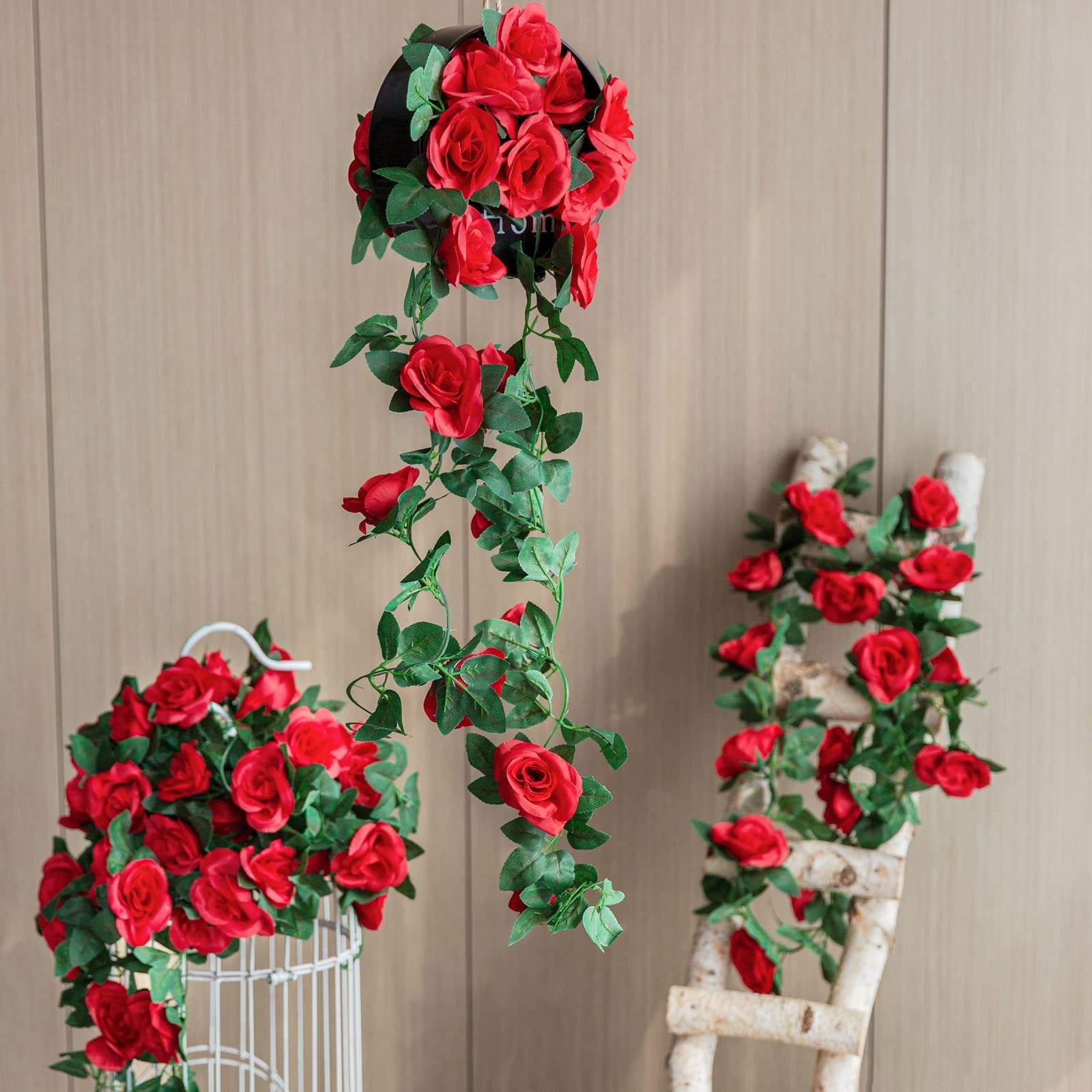 Artificial red rose garlands with green leaves draped on a white birdcage and a birch ladder, and hanging from a black planter.