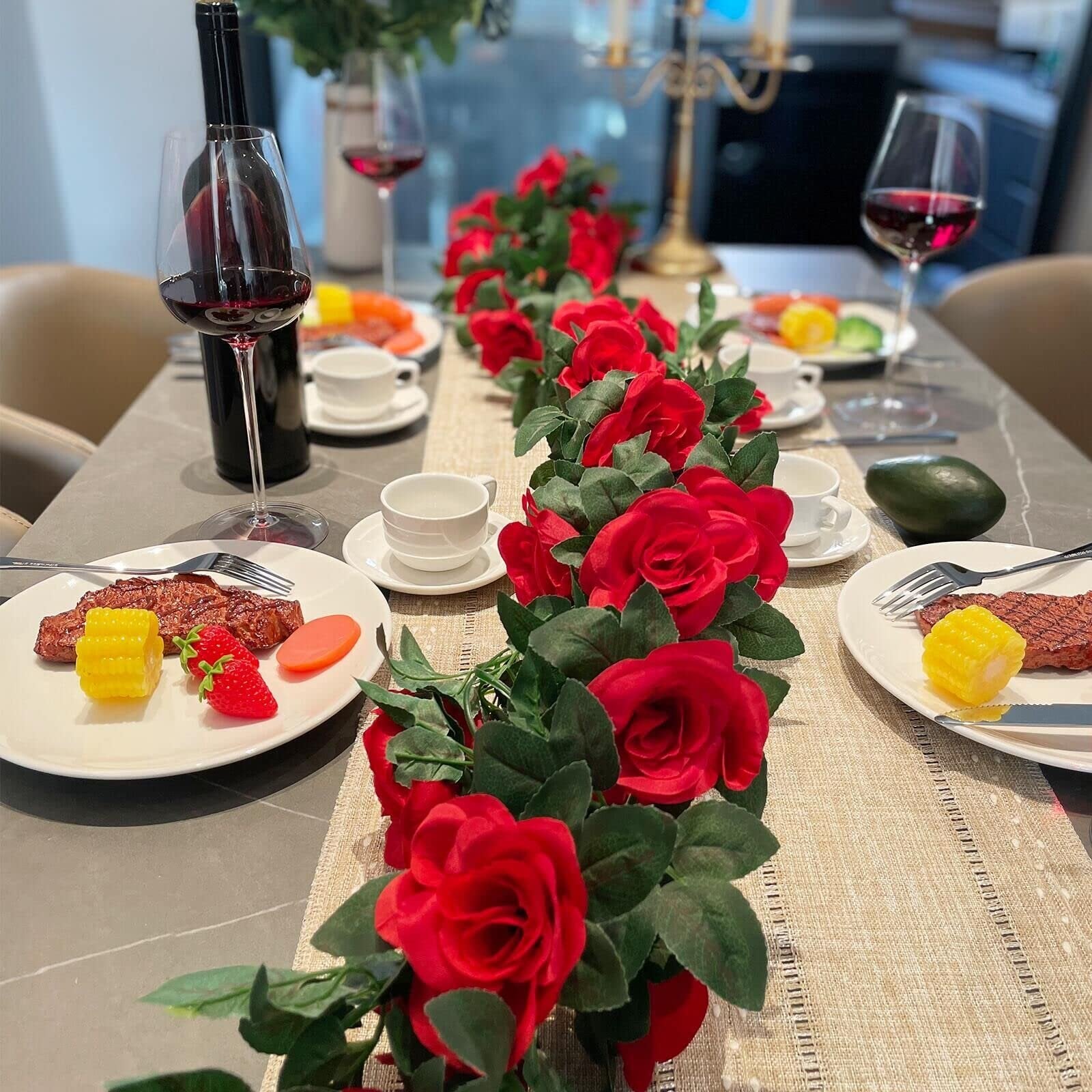 Artificial red rose garland draped across a dining table set for a meal with wine and steaks.