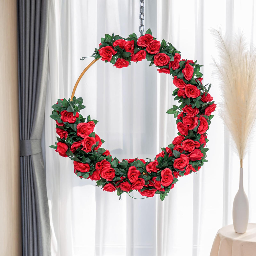 Circular artificial red rose garland with green leaves hanging from a chain, against a backdrop of sheer curtains and a vase with pampas grass.