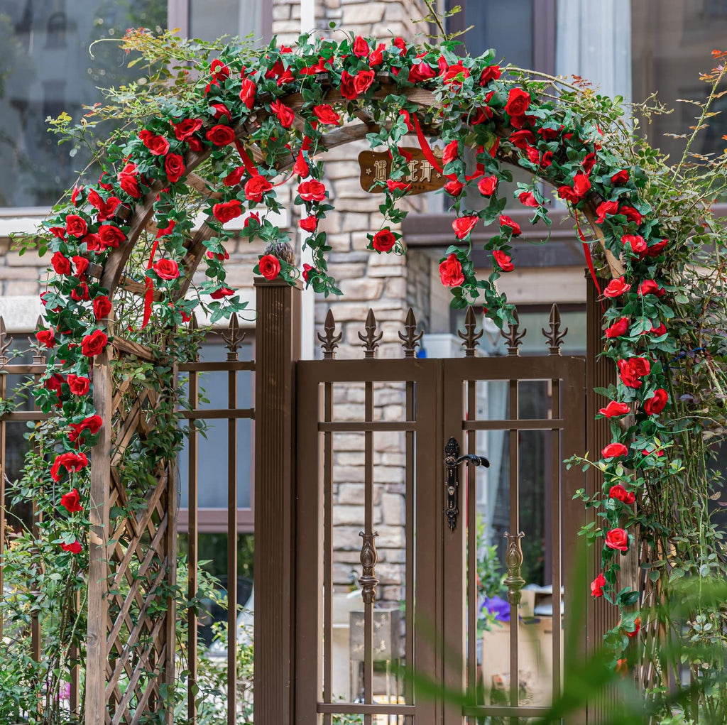 Artificial red rose vine garland draped over a wooden archway and gate, with green leaves and red ribbons.