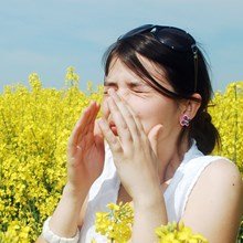 Woman with eyes closed and hands over her face, suffering from allergies in a field of yellow flowers.