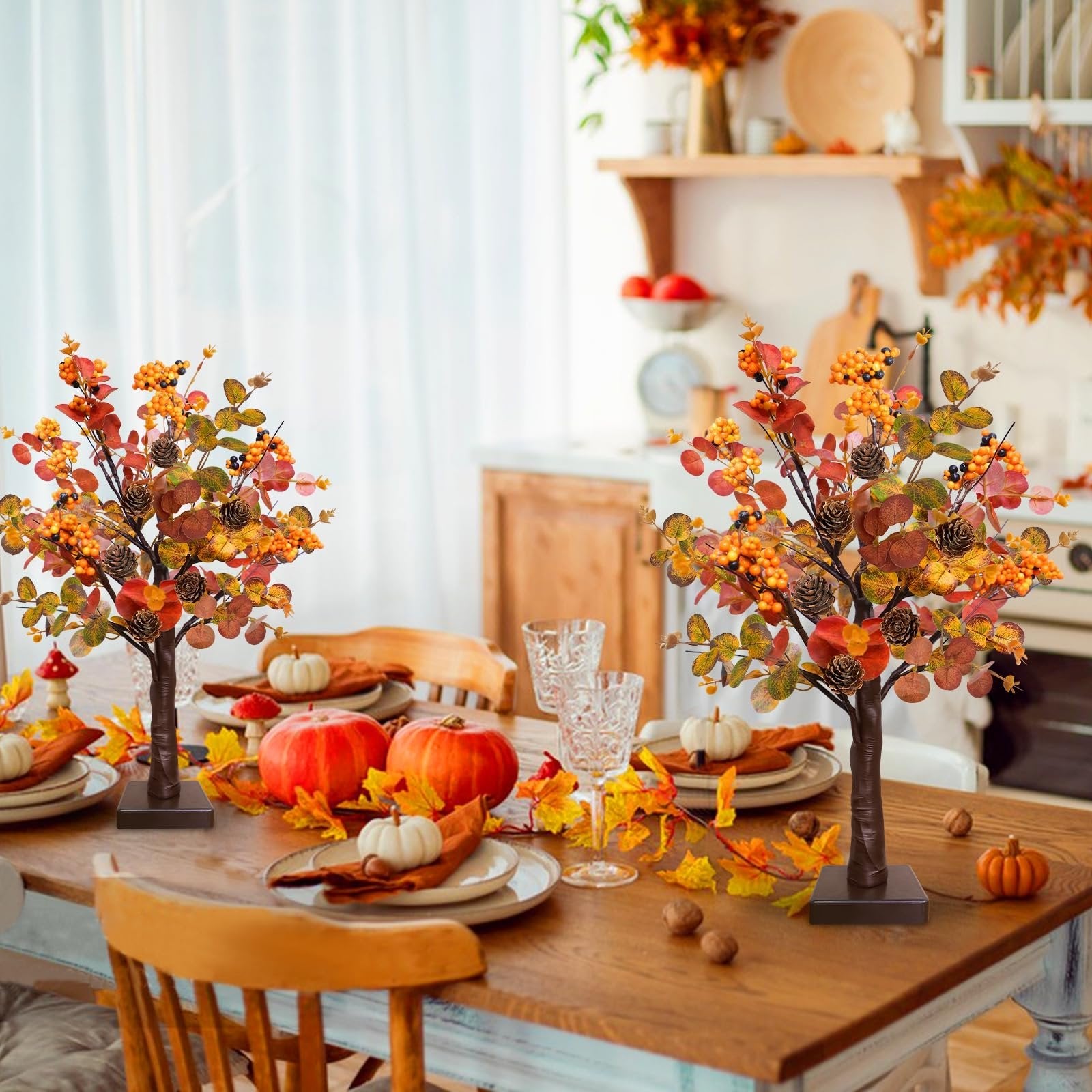 Two prelit fall tabletop trees with warm LEDs, eucalyptus leaves, pine cones, and berries on a dining table set for fall.