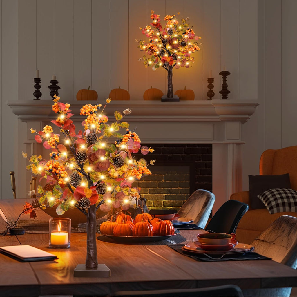 Two prelit fall tabletop trees with warm LEDs, eucalyptus leaves, pine cones, and berries on a dining table with pumpkins and a candle.