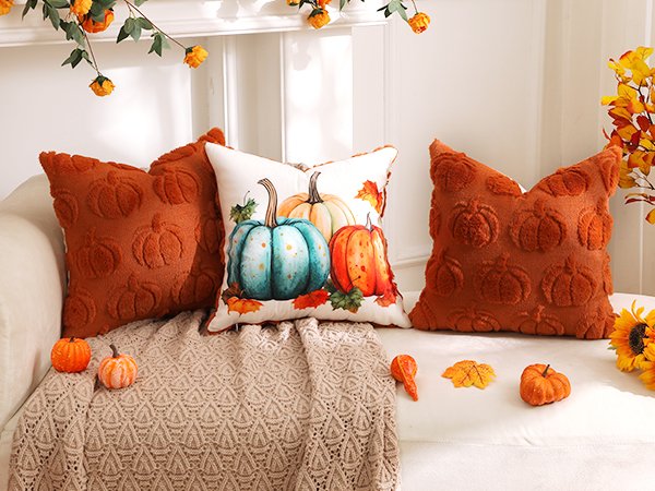 Two rust orange faux fur pumpkin throw pillow covers and one white cover with painted pumpkins on a couch with a blanket.