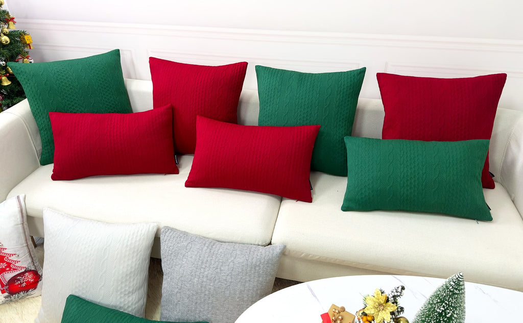 Several red and green knit textured pillow covers arranged on a white sofa, with a Christmas tree visible on the left.