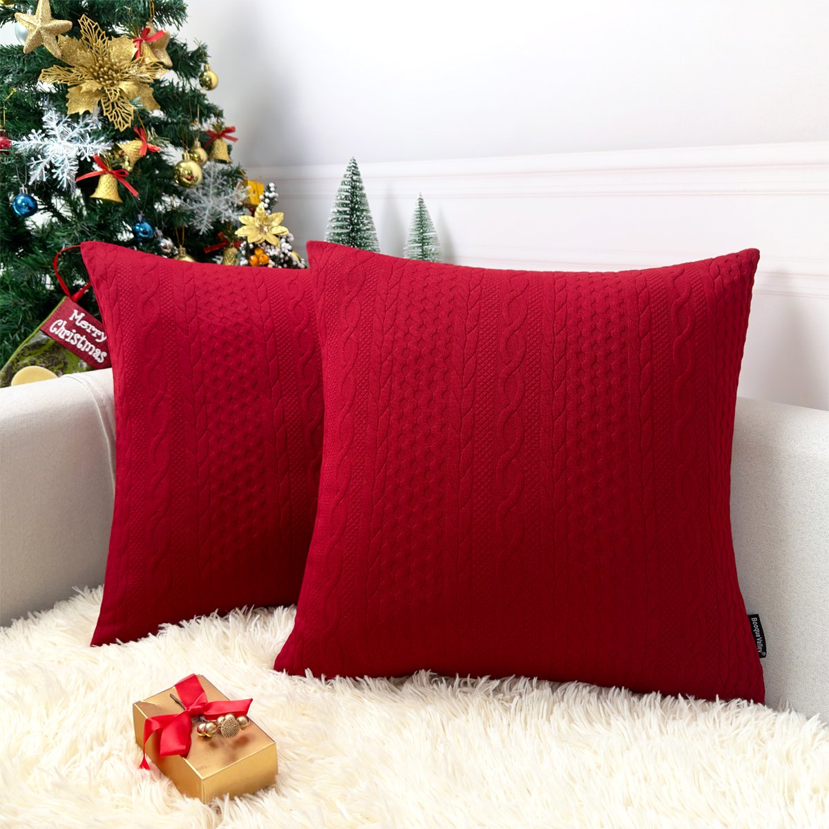 Two red knit Christmas pillow covers with embossed cable texture on a couch with a Christmas tree and gift.