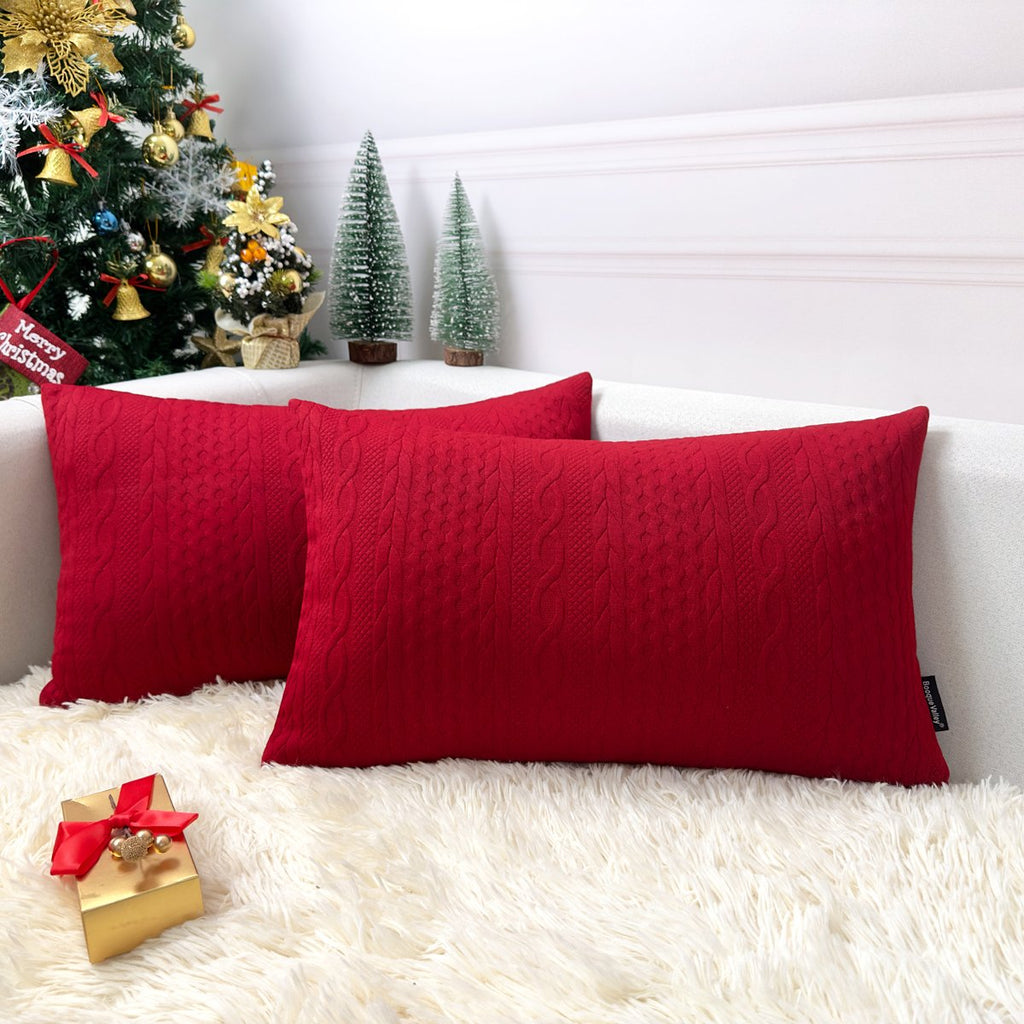 Two red knit Christmas pillow covers with cable knit texture on a white couch with a Christmas tree and gift.