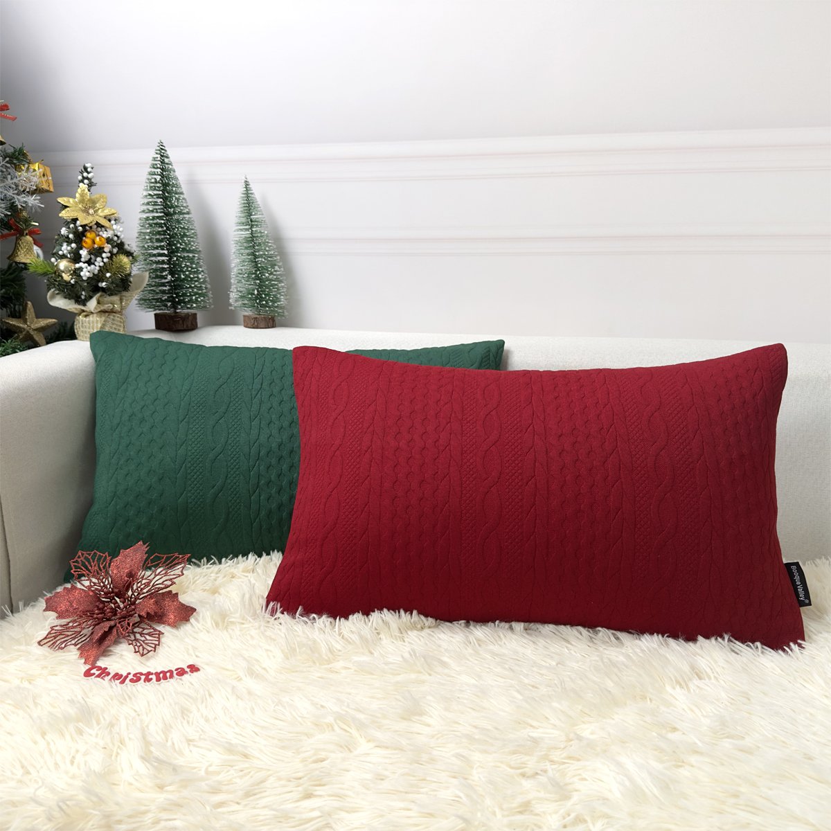 Two 18x18 inch red and green cable knit Christmas pillow covers on a sofa with decorative trees.
