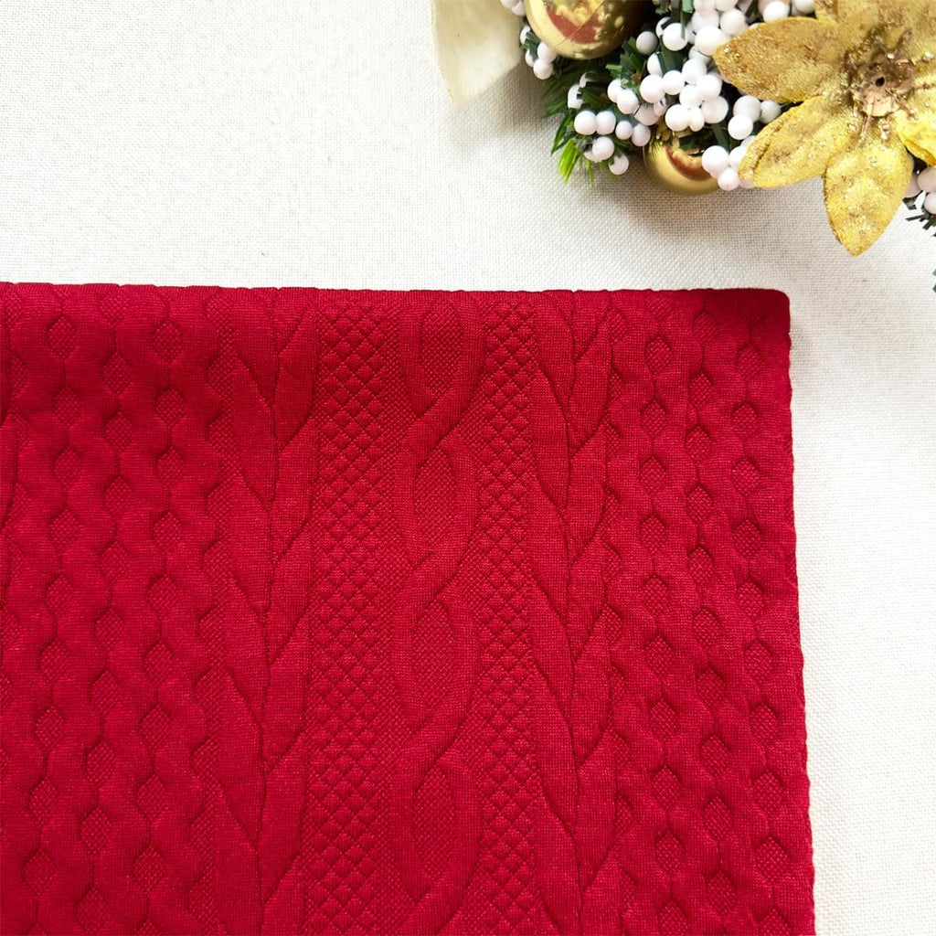 Red Christmas pillow cover with embossed knit texture, shown flat with festive decor in the upper right corner.