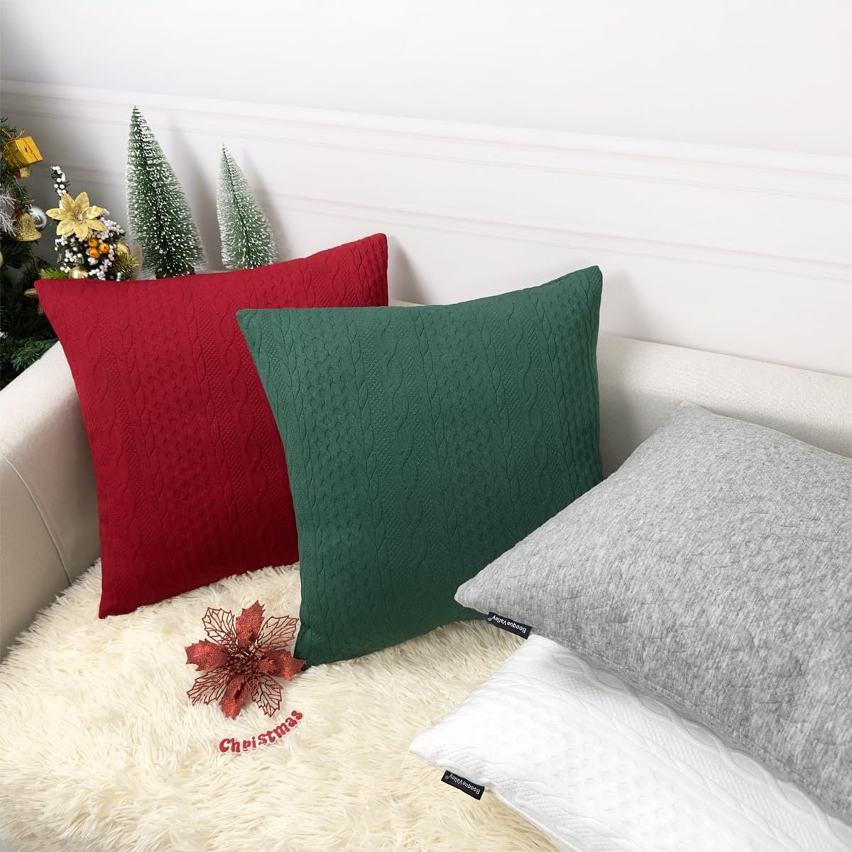 Three Christmas-themed knit pillow covers in red, green, and grey arranged on a sofa with a fluffy white throw.