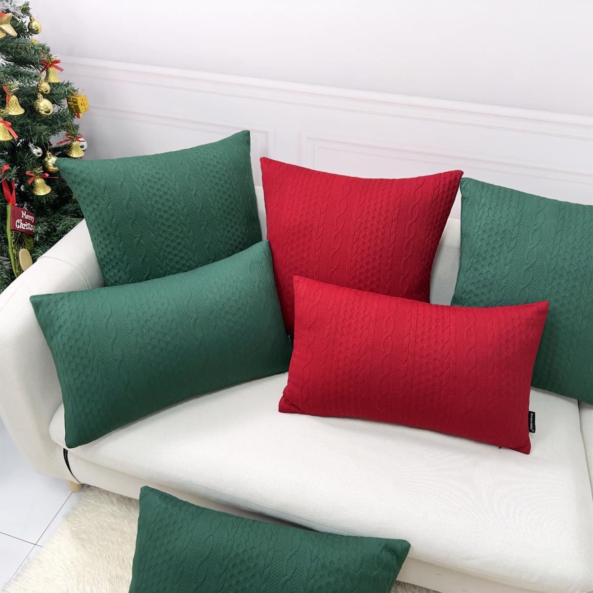 Christmas pillow covers in red and green with cable knit texture arranged on a white sofa next to a decorated Christmas tree.