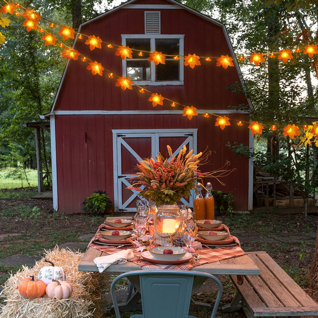 Outdoor dining table set with fall decor and maple leaf string lights draped over a red barn
