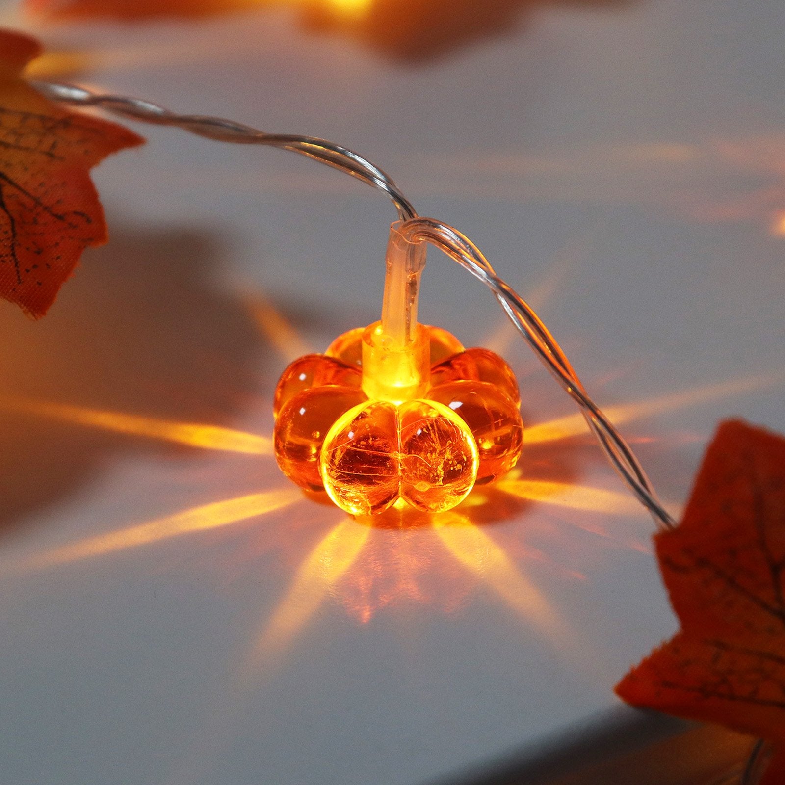 Close-up of a lit pumpkin-shaped LED light on a string with artificial maple leaves, casting warm orange light.