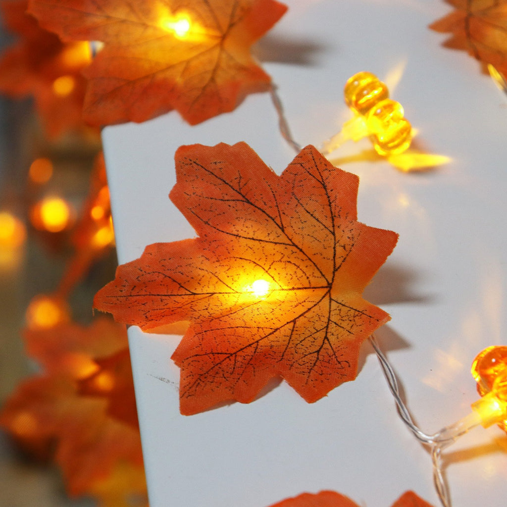 Close-up of illuminated artificial maple leaves and pumpkin-shaped lights on a string, set against a white surface.