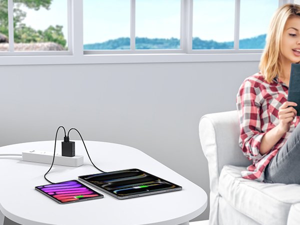 Dual USB C wall charger block plugged into power strip, charging two tablets on a white table, woman in background reading