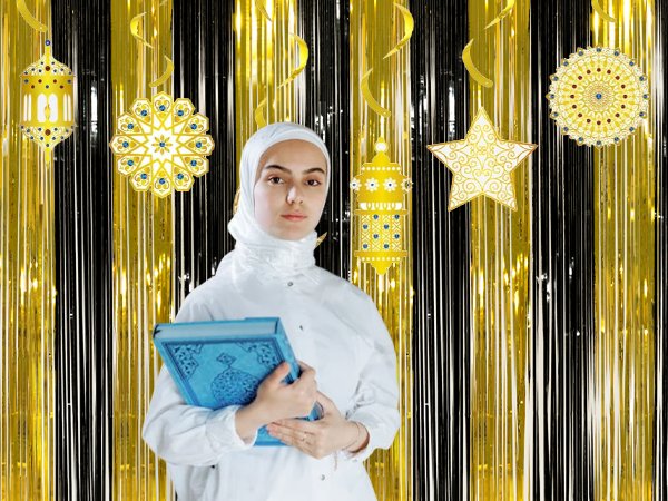 Woman in white hijab holding a blue book in front of a black and gold fringe curtain backdrop with decorative lanterns and stars