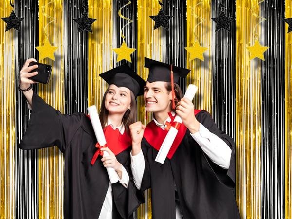 Two graduates in caps and gowns taking a selfie in front of a black and gold fringe curtain backdrop with hanging stars.