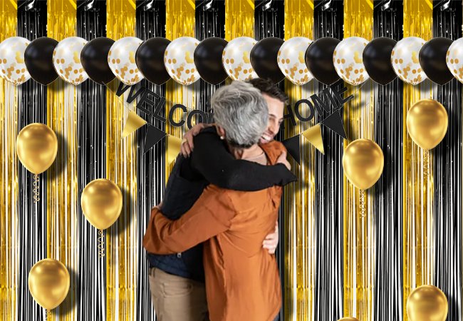 Two people hugging in front of a black and gold fringe curtain backdrop with balloons and a welcome home banner.