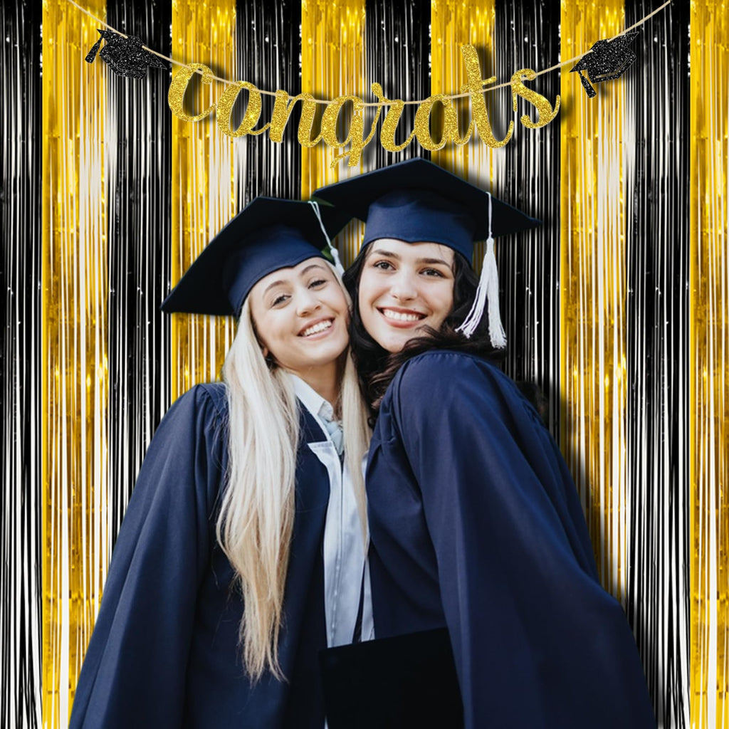 Two smiling graduates in caps and gowns pose in front of a black and gold fringe backdrop with a 'congrats' banner.