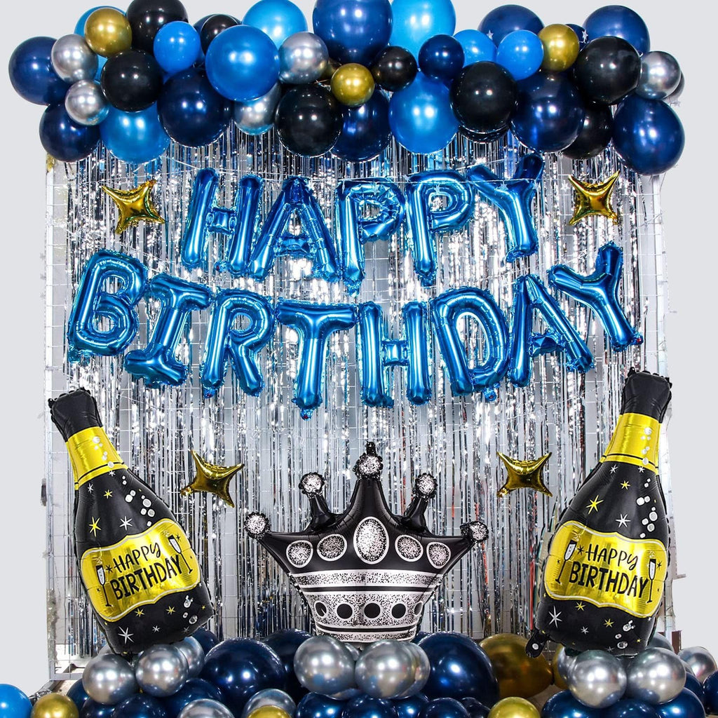 Birthday party backdrop with blue, black, and silver balloons, foil fringe curtain, and foil balloons shaped like champagne bottles and a crown.