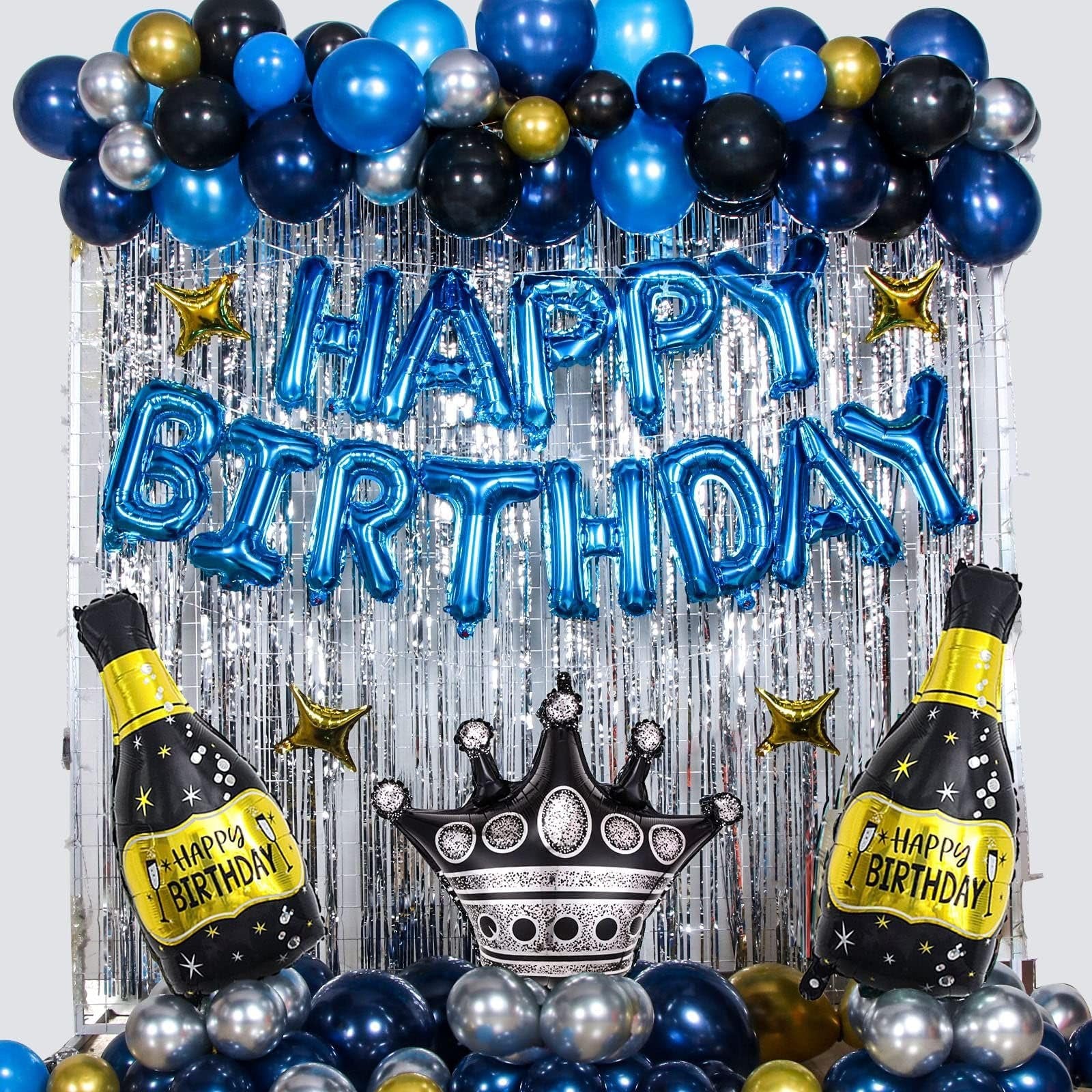 Birthday party backdrop with blue, black, and silver balloons, foil fringe curtain, and foil balloons shaped like champagne bottles and a crown.