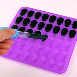 Purple silicone candy mold with 40 skull cavities being filled with dark liquid using a blue dropper